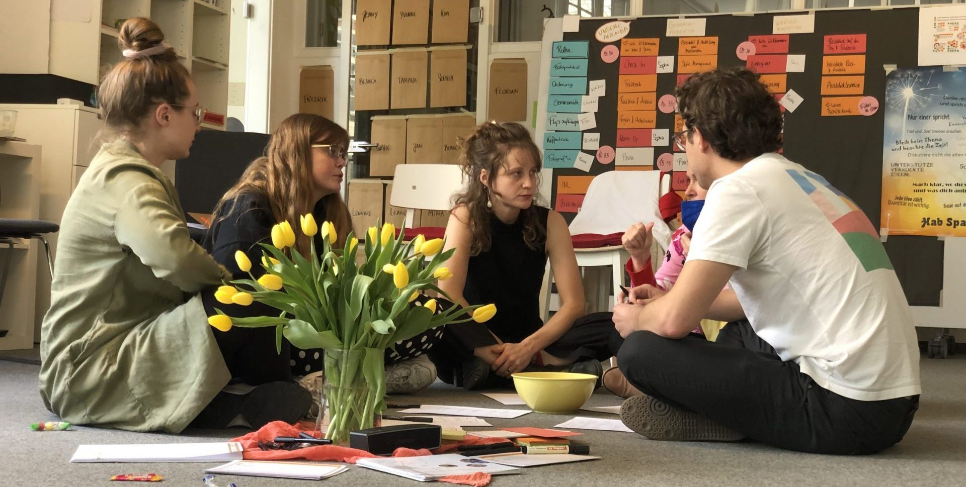 5 junge Menschen sitzen in einem Workshopraum vor einem Blumenstrauß auf dem Boden